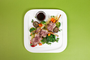 Baked lamb with vegetables on a white platter with red sauce top view on a green background