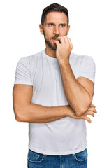 Handsome man with beard wearing casual white t shirt looking stressed and nervous with hands on mouth biting nails. anxiety problem.