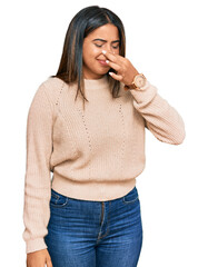 Young latin girl wearing wool winter sweater smelling something stinky and disgusting, intolerable...