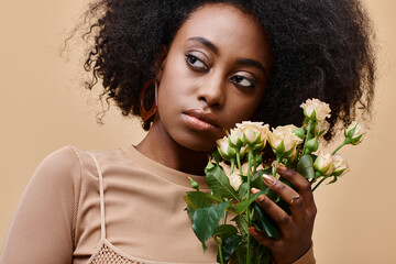 pensive curly african american woman holding tiny roses on beige background, peach fuzz color