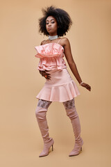 fancy african american woman in 20s, posing in peach ruffle top and trendy over-knee boots on beige