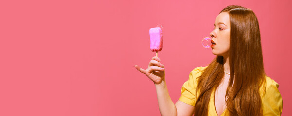 Yong redhead woman holding fake ice cream made of soap and making soap bubble with mouth against pink background. Concept of pop art photography, creativity. Banner. Empty space for ad