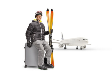 Young man with a pair of skiis sitting on a suitcase in front of an airplane