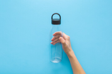 Young adult woman hand holding and showing new empty transparent plastic bottle with black carry lid on light blue table background. Pastel color. Closeup. Top down view.