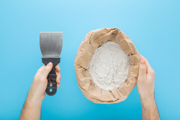 Man hands holding spatula and opened paper bag of white dry finishing putty for plastering ceiling or wall surfaces on blue background. Repair work of home. Closeup. Point of view shot. Top down view.