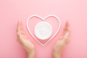 Young adult woman hands showing white heart shape with cream jar on light pink table background. Pastel color. Care about clean and soft body skin. Closeup. Point of view shot. Top down view.