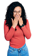 Young hispanic woman with curly hair wearing glasses laughing and embarrassed giggle covering mouth...