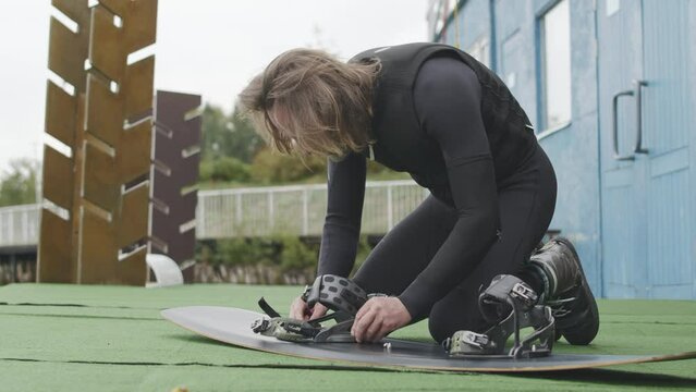 Long-haired Caucasian male surfer in wetsuit setting up wakeboard bindings and adjusting wakeboard stance width while preparing for ride in wake park