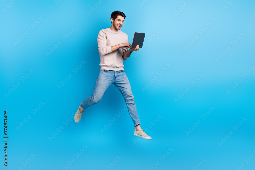 Poster full size photo of nice young man jump use netbook empty space wear beige sweatshirt isolated on blu