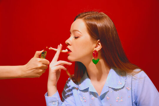 Young redhead woman with red lips smoking doll part against red background. Smoking for fun, quitting bad habit. Concept of pop art photography, creativity, surrealism, weirdness