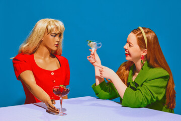 Fake friends. Young redhead positive woman smiling, drinking cocktail and talking to mannequin against blue background. Concept of pop art photography, creativity, psychology, fantasy