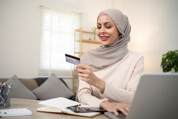 Young happy muslim woman in hijab at home using laptop shopping online with credit card while sitting on desk