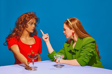 Young redhead woman drinking cocktail, talking, arguing with mannequin against blue background. Intense communication. Concept of pop art photography, creativity, surrealism, psychology
