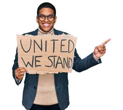 Young handsome hispanic man holding united we stand banner smiling happy pointing with hand and finger to the side