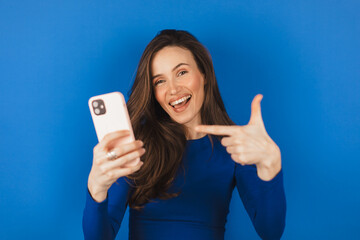 Excited brunette girl wear blue top smiling amazed and pointing at smartphone screen isolated on...