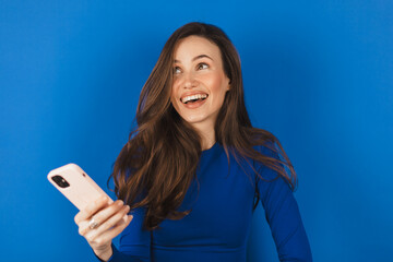 Overjoyed happy excited woman say wow, glad to receive text message informing about salary, rejoices good news, stares at mobile phone, gestures actively from happiness isolated blue background.