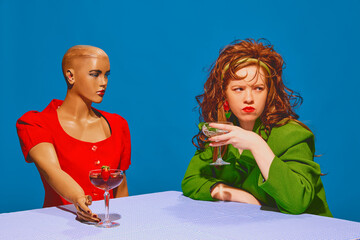Young redhead woman with messy hair drinking cocktail, sitting by table with bald mannequin against blue background. Thoughtful talk. Concept of pop art photography, creativity, psychology