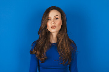Fototapeta premium Portrait of caucasian woman with no expression. ID or passport photo full collection of diverse face and expressions. Calm interesting woman in blue long sleeves with serious face on blue background. 