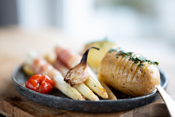 Fried white asparagus wrapped in bacon with herbed potatoes and tomatoes on black ceramic plate Close-up with short depth of field. - 774881261