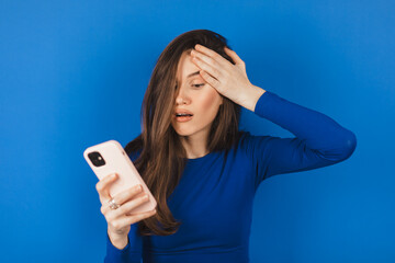 Mad and shock brunette woman make facepalm using mobile phone isolated on blue background. Yelling...