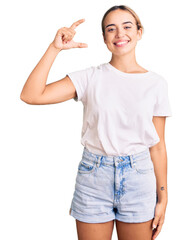 Young beautiful blonde woman wearing casual white tshirt smiling and confident gesturing with hand doing small size sign with fingers looking and the camera. measure concept.