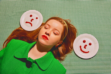 Young redhead woman lying with paper plates showing happy and sad faces. Choosing mood for today....
