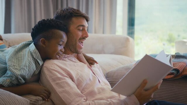 Father in multi-racial family sitting with son on sofa at home reading book  together-- shot in slow motion