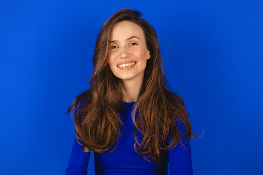 Happy cheerful young woman wearing blue long sleeves top rejoicing at positive news or birthday gift, looking at camera with joyful and charming smile. Brunette student girl relaxing isolated blue.
