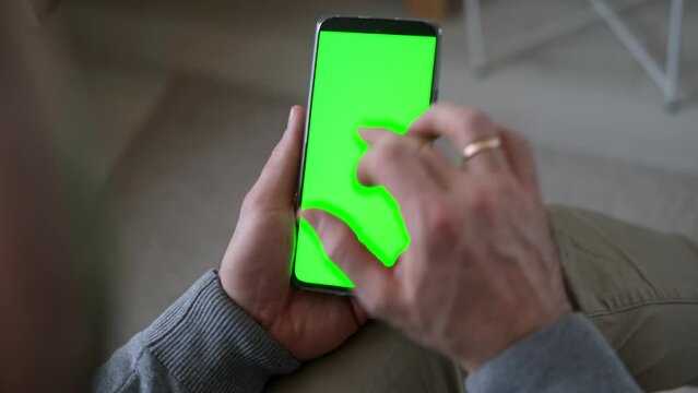 Young man sitting at home holding smartphone green mock-up screen in hand. Female person use chroma key mobile phone. Vertical mode. Touching, swiping display, tapping, surfing internet social media