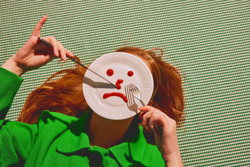 Young redhead woman lying with knife and fork over paper plates showing sad face. Choosing mood for today. Concept of pop art photography, creativity, psychology, surrealism
