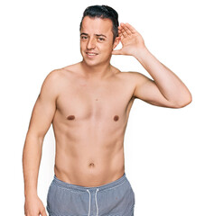 Fototapeta premium Handsome young man wearing swimwear shirtless smiling with hand over ear listening an hearing to rumor or gossip. deafness concept.