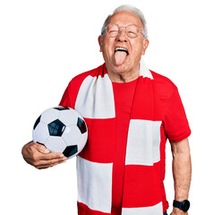Senior man with grey hair football hooligan holding ball sticking tongue out happy with funny...