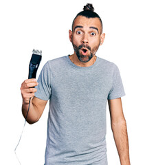 Hispanic man with ponytail holding electric razor machine scared and amazed with open mouth for...