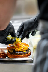 A person in gloves is meticulously assembling a burger, adding pickles on top of the melted cheese and grilled patty