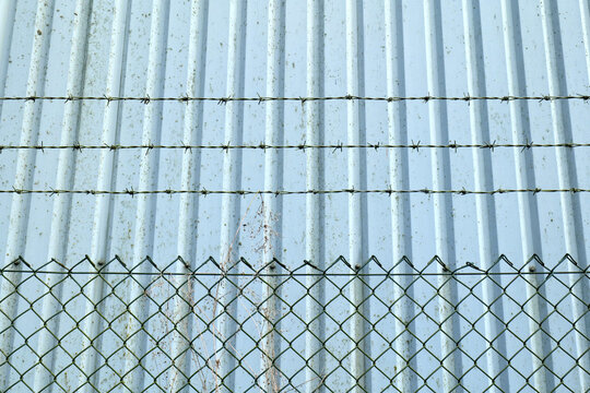 Barbed wire metal fence provides security protecting pale industrial unit wall.