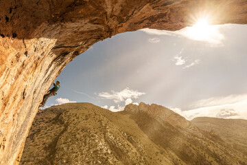 A man is climbing a rock face with a sun shining on him. The sun is in the background and the...