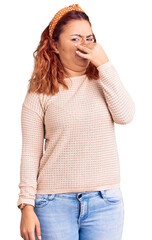 Fototapeta premium Young latin woman wearing casual clothes and diadem smelling something stinky and disgusting, intolerable smell, holding breath with fingers on nose. bad smell