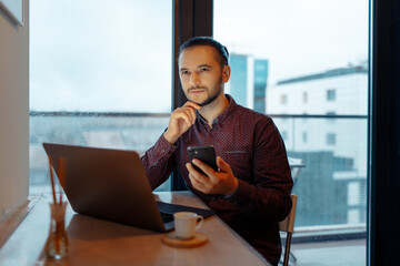 Portrait of thoughtful man working at laptop with smartphone in hand, background of panoramic windows.