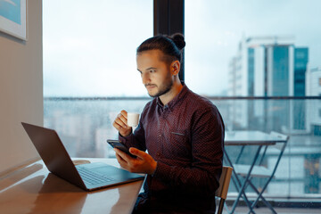 Portrait of handsome man working at laptop, holding smartphone and cup of espresso coffee in hands, background of panoramic windows.