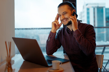Portrait of happy man working at laptop, talking on smartphone, background of panoramic windows.