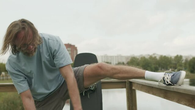 Medium shot of Caucasian mature wake surfer stretching his legs on pier, warming up before wakeboarding on water