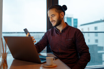 Portrait of handsome smiling man looking in smartphone while working at laptop, background of panoramic windows.