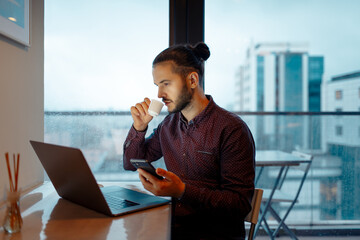 Portrait of handsome man, drinking coffee espresso, working at laptop, holding smartphone in hand, background of panoramic windows.
