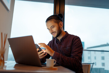 Portrait of smiling handsome man working at laptop, using smartphone, background of panoramic windows.