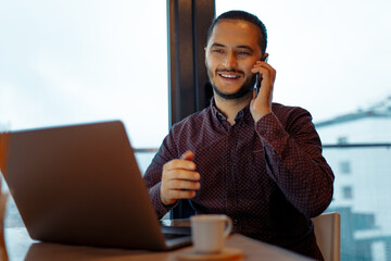 Portrait of smiling handsome man working at laptop, talking on smartphone, background of panoramic windows.