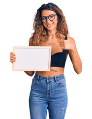 Young hispanic woman with tattoo holding empty white chalkboard smiling happy and positive, thumb up doing excellent and approval sign