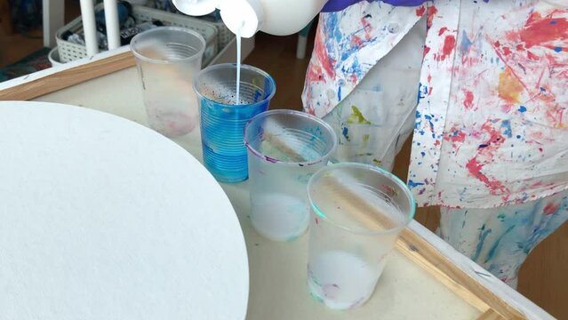 Female artist mixing acrylic paint. Close up on hands of a female artist wearing violet rubber gloves and colorful stained shirt pouring white acrylic paint from big bottle to plastic cups. 