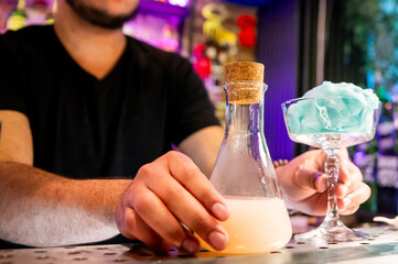 A man holds a flask with a cork stopper, next to a glass emitting blue smoke in a bar setting.