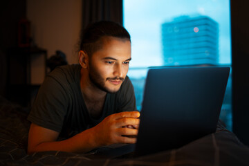 Portrait of young serious man working at laptop while lying on the bed close to evening time.