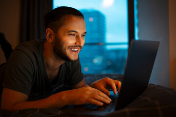 Portrait of young smiling man typing on keyboard of laptop while lying on the bed close to evening time.